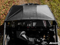 Overhead view of a black UTV with the Can-Am Maverick X3 Primal Soft Top Roof parked on grass and fallen leaves, showing its angular dashboard and roll cage. Sunlight casts shadows across the roof, with the SuperATV logo in the lower right corner.