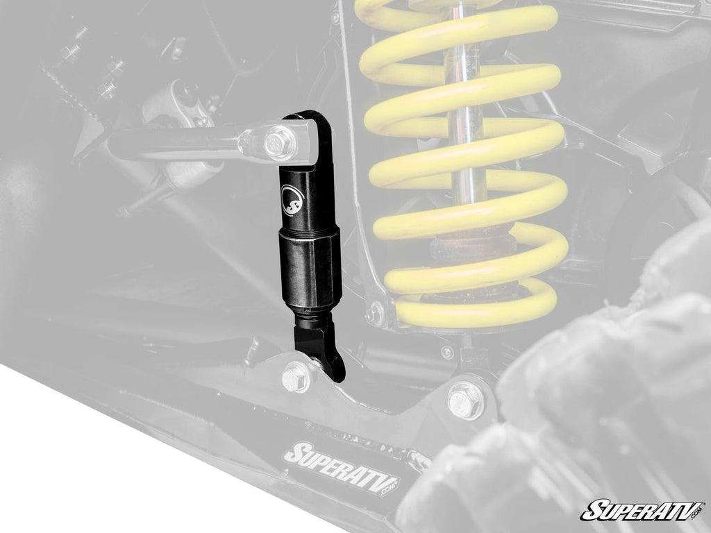 A close-up of a Can-Am Maverick X3 Sway Bar Shock installed on an off-road vehicle’s suspension, with a yellow coil spring and background components blurred. The SuperATV logo appears near the lower right corner.