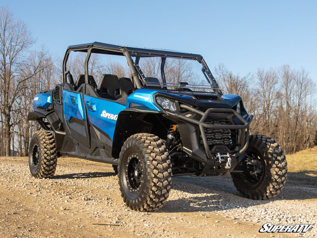 A blue off-road utility vehicle equipped with Can-Am Commander 1000 Atlas Pro 1.5 Forward Offset A-Arms and rugged tires is parked on a dirt path, featuring an open cabin and roll cage. Leafless trees and dry grass are in the background under a clear sky.