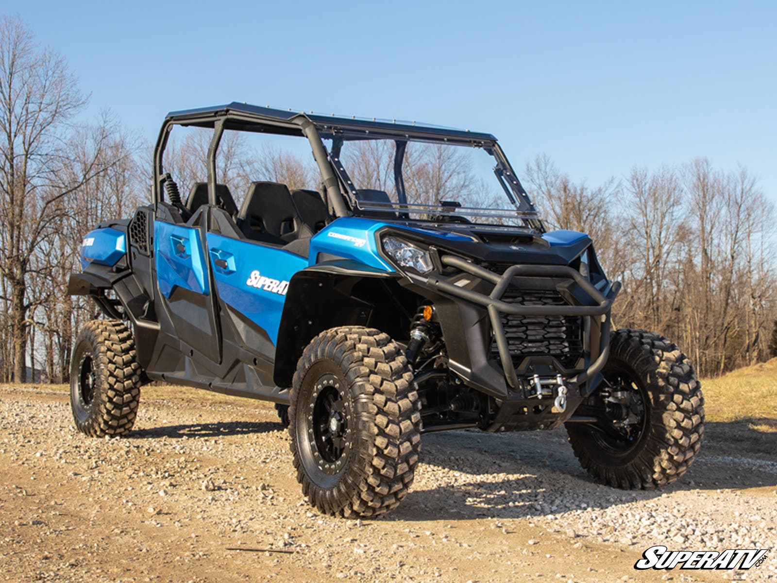A blue off-road utility vehicle equipped with Can-Am Commander 1000 Atlas Pro 1.5 Forward Offset A-Arms and rugged tires is parked on a dirt path, featuring an open cabin and roll cage. Leafless trees and dry grass are in the background under a clear sky.