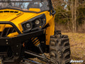 Close-up of a rugged yellow off-road vehicle’s front left side, featuring a large muddy tire, headlight, suspension system, and grill. Trees and leaf-strewn grass are in the background. Can-Am Commander Deluxe Plug & Play Turn Signal Kit shown.