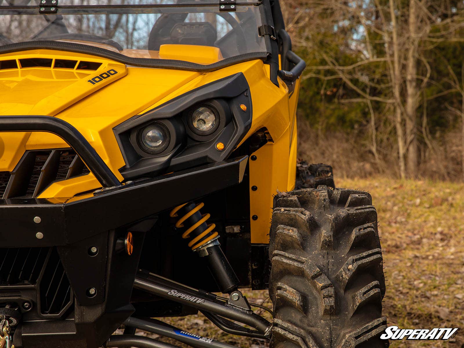 Close-up of a rugged yellow off-road vehicle’s front left side, featuring a large muddy tire, headlight, suspension system, and grill. Trees and leaf-strewn grass are in the background. Can-Am Commander Deluxe Plug & Play Turn Signal Kit shown.