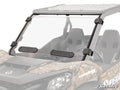 A close-up of the Can-Am Maverick Sport Vented Full Windshield shows the camouflage-patterned off-road vehicle with a clear windshield secured by four black brackets and two rectangular black vents at the bottom, set against a plain white background.