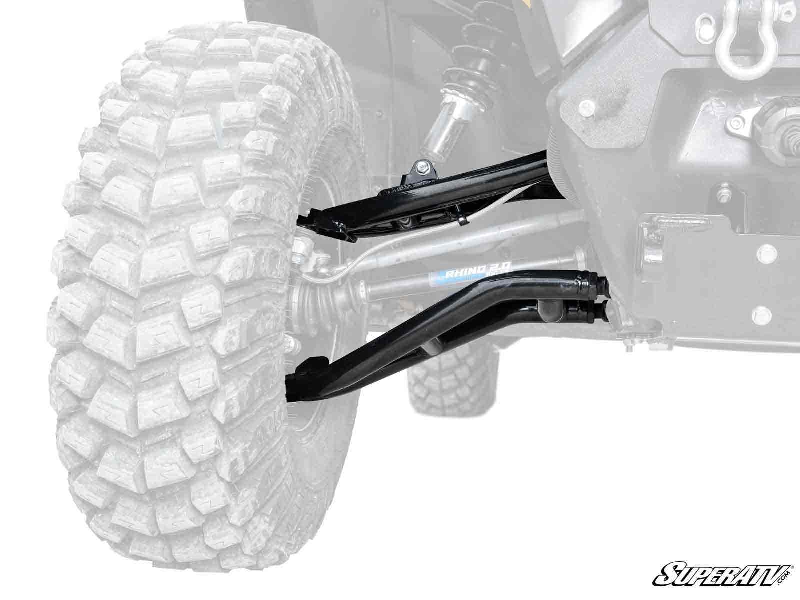 Close-up of a Can-Am Defender HD10 Atlas Pro 2 Forward Offset A-Arms on the front left wheel, with visible black suspension components and rugged knobby tire tread; control arms and axle shown in sharp detail against a faded vehicle backdrop.
