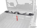 A Can-Am Defender Bed Winch, equipped with a winch and red strap, is mounted at the front of a utility vehicle’s cargo bed. The faded bed and cab interior highlight the winch mount. SuperATV logo is shown in the bottom right corner.