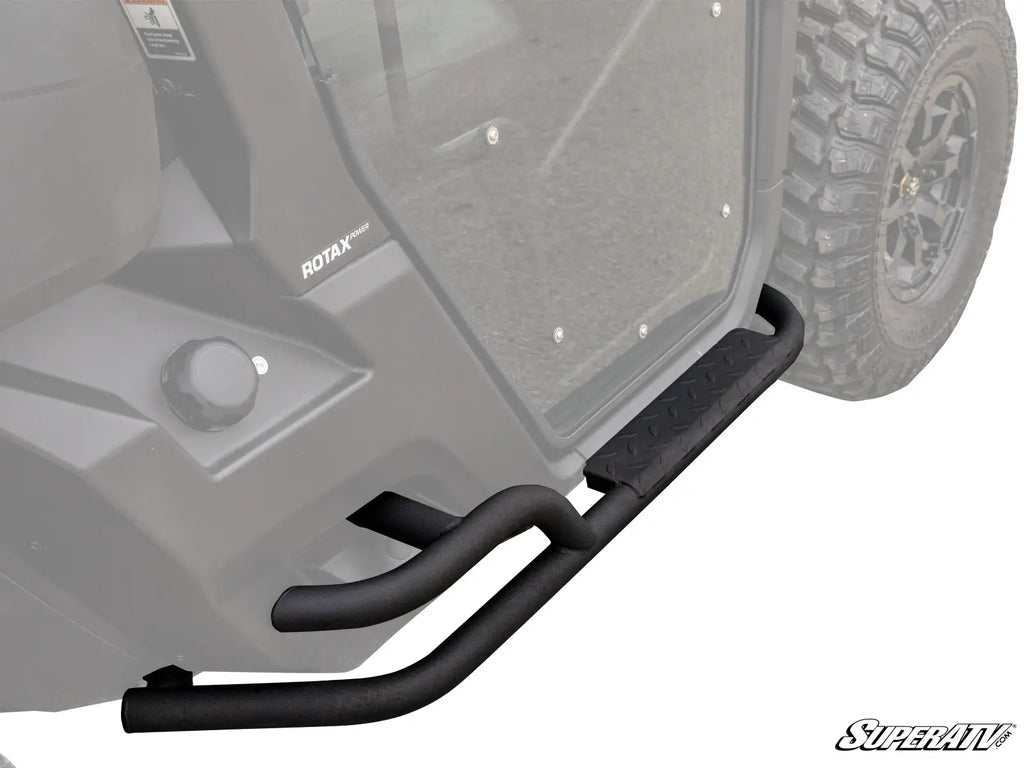 Close-up of Can-Am Defender Heavy Duty Nerf Bars, black tubular metal with an angular step plate, installed on an off-road vehicle showing a ROTAX label, clear door, and rugged tire.