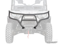 The Can-Am Defender Front Bumper, a black, heavy-duty bumper with tubing and a central protective plate, is mounted on an off-road vehicle. It has a winch opening for rugged terrain. The SuperATV logo appears in the lower right corner.