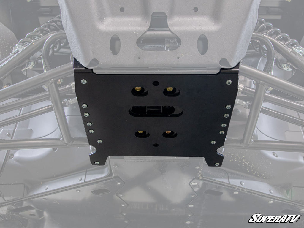 Close-up view of a Can-Am Defender Frame Stiffener, a black metal plate mounted under an off-road vehicle chassis with bolts securing it to the frame and suspension. The faint SuperATV logo appears in the images bottom right corner.