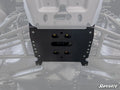 Close-up view of a Can-Am Defender Frame Stiffener, a black metal plate mounted under an off-road vehicle chassis with bolts securing it to the frame and suspension. The faint SuperATV logo appears in the images bottom right corner.