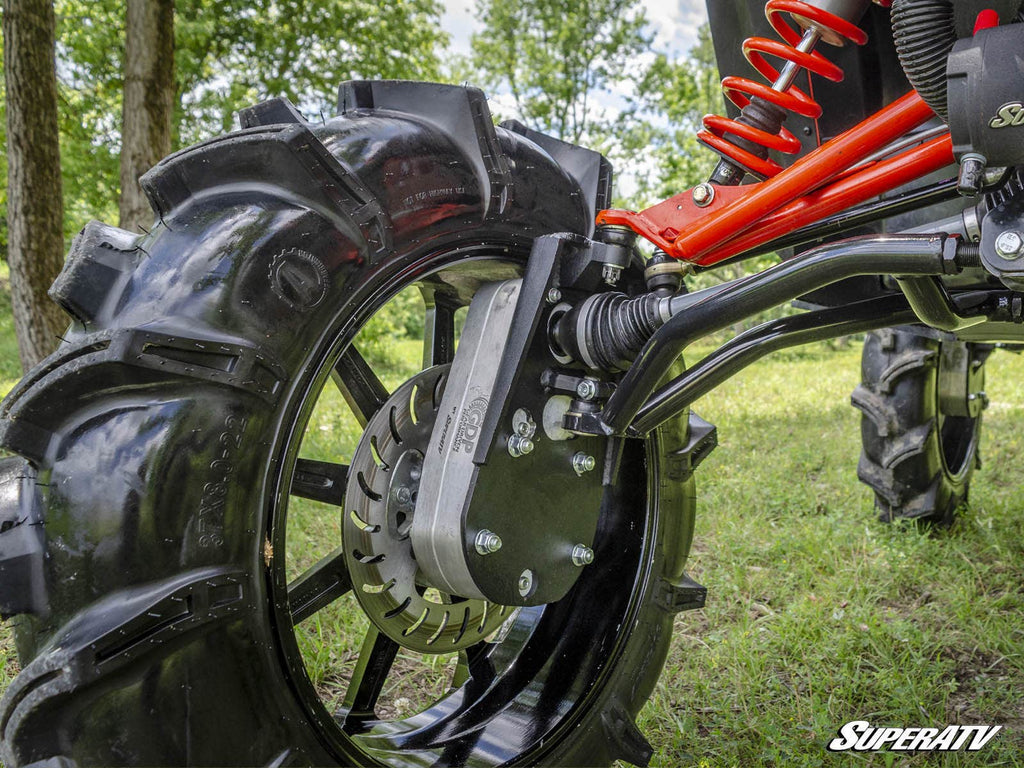 Close-up of a Can-Am Maverick 8 Portal Gear Lift, featuring a large off-road tire with deep treads on a metallic wheel. The visible suspension and disc brake stand out against grass and sunlight, with the SuperATV logo in the corner.
