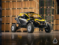An Assault Industries Can-Am Maverick R High-Clearance Boxed A-Arms vehicle, in yellow and black, is parked indoors on a shiny concrete floor among stacks of cardboard boxes, lit by industrial lights with a geometric white logo in the corner.