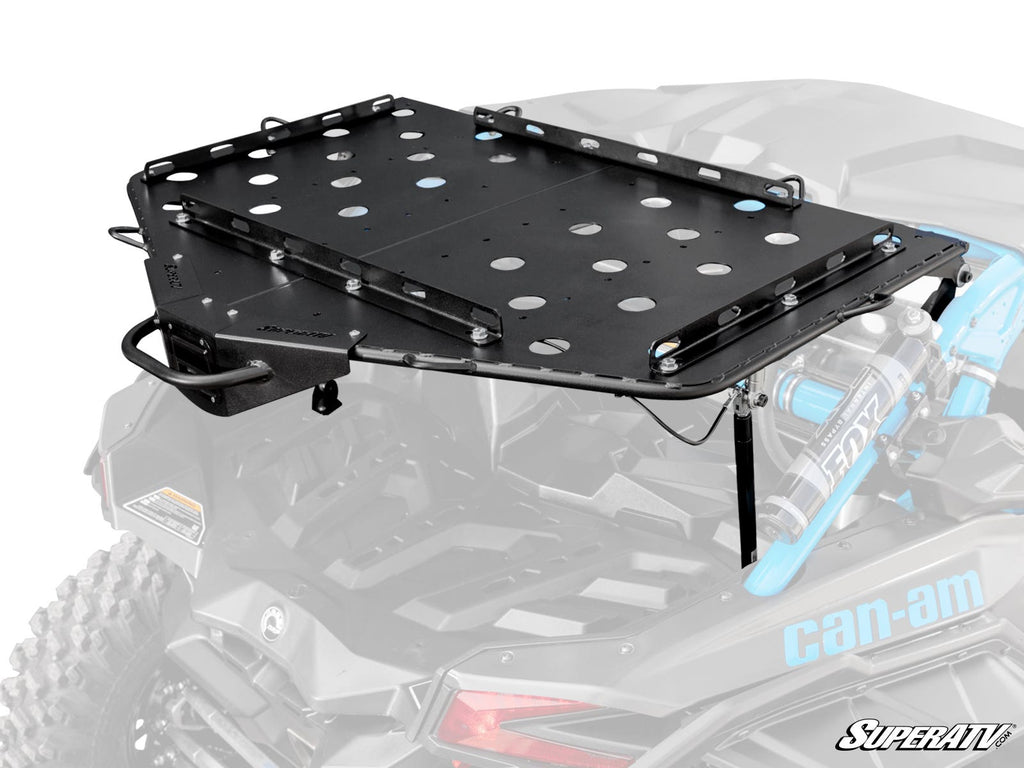 The Can-Am Maverick X3 Cargo Rack Alpha, a black metal rack with circular cutouts, is mounted on the rear of a Can-Am off-road vehicle. It features side supports, attachment points, and the SuperATV logo with visible rear suspension below.