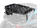 The Can-Am Maverick X3 Cargo Rack Alpha, a black metal rack with circular cutouts, is mounted on the rear of a Can-Am off-road vehicle. It features side supports, attachment points, and the SuperATV logo with visible rear suspension below.