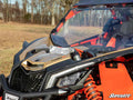 A close-up of a gold and black off-road Can-Am Maverick X3 shows its Billet Shock Tower Brace, suspension, windshield, and “Turbo R” branding. Parked on grass with leafless trees and blue sky, the SuperATV logo is at the bottom right.