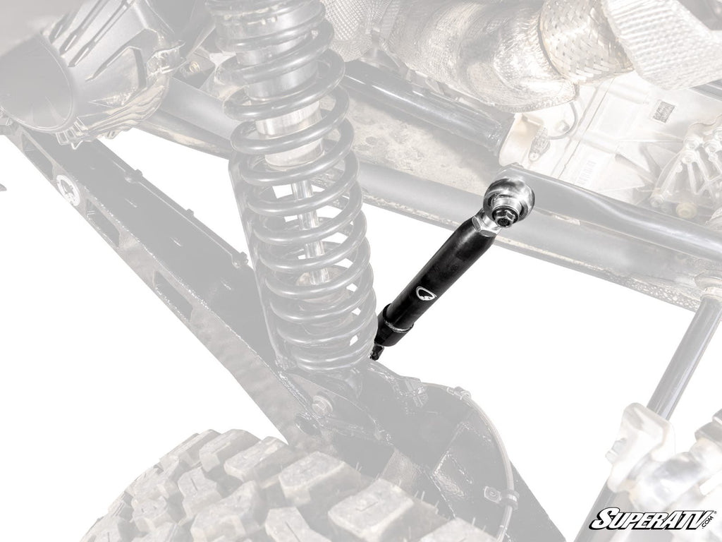 Close-up of a Can-Am Maverick R Sway Bar Shock with a large coil spring, black shock absorber, and rugged tire. Mostly black and gray parts; “SuperATV” logo at bottom right. Some areas are desaturated for emphasis.