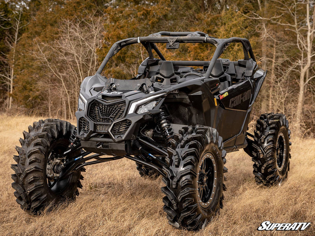 A black Can-Am Maverick X3 with a 6 Lift Kit and large rugged tires is parked on dry grass before leafless trees. Its angular design, roll cage, and the SuperATV logo in the bottom right are visible.