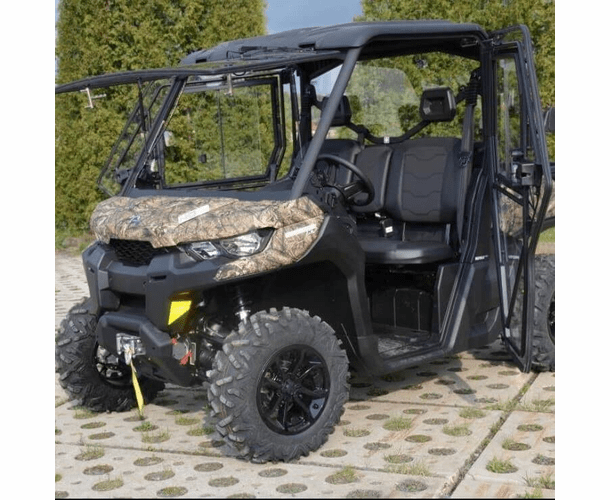 A Can Am Defender Full Hard Cab Enclosure UTV with camo finish and large off-road tires is parked on pavement. It features a partially open windshield, mesh side doors, black seats, a front winch, and bushes in the background.
