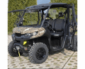 A Can Am Defender Full Hard Cab Enclosure UTV with camo finish and large off-road tires is parked on pavement. It features a partially open windshield, mesh side doors, black seats, a front winch, and bushes in the background.