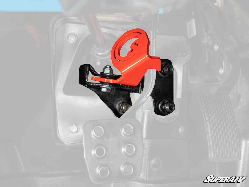 A bright red and black Can-Am Maverick X3 Brake Lock is mounted on a metal base with bolts inside a vehicle. The background is partially faded, drawing focus to the brake lock. The SuperATV logo appears in the bottom right corner.