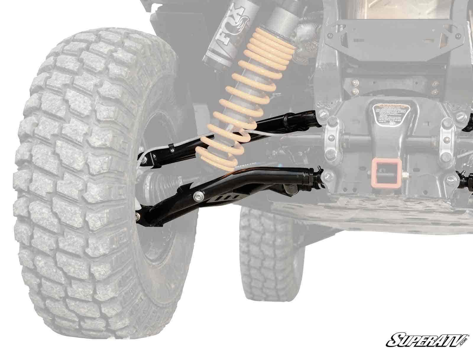 Close-up of a Can-Am Maverick Sport Atlas Pro Rear Offset A-Arms suspension, featuring a large knobby tire, coil-over shock, and durable black control arms. The background is faded; “SuperATV” logo appears in the bottom right corner.