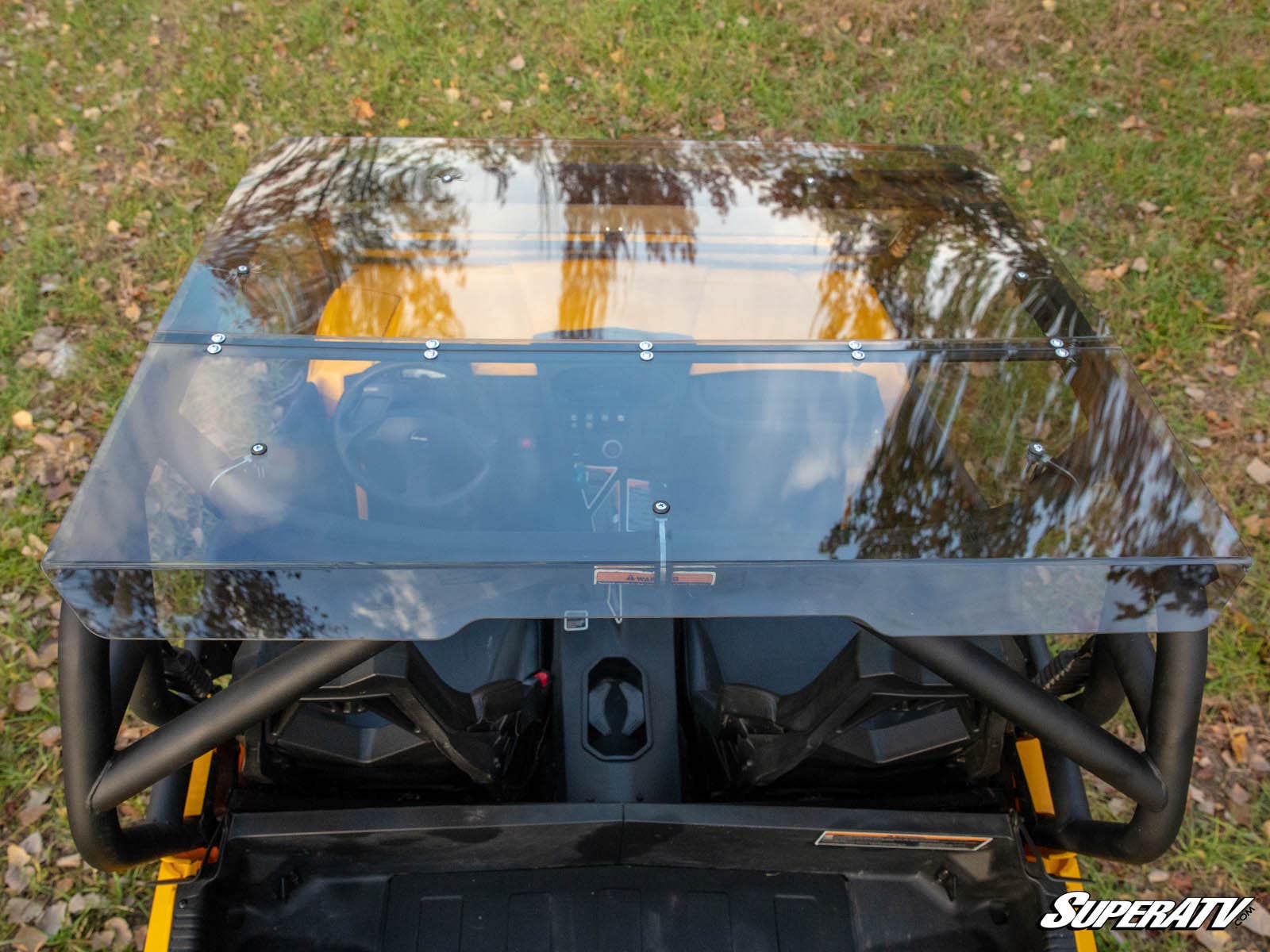 Overhead view of a yellow off-road vehicle fitted with a Can-Am Commander Tinted Roof reflecting trees. The interior has two bucket seats and a minimalist dashboard. Vehicle is parked on grass with fallen leaves; SuperATV logo in the corner.