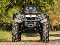 A beige and black ATV fitted with Can-Am Outlander (2019+ Gen 2) High Clearance 2 Forward Offset A-Arms faces forward on a dirt path, showcasing large muddy tires and robust suspension, with green trees and blurred foliage in the background.