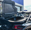 A close-up shows the open Cargo Box for all Can-Am X3 UTVs on a black utility vehicle, with the lid raised to reveal an empty compartment. The vehicle is parked outside a modern blue building, and another vehicle is partially visible behind it.