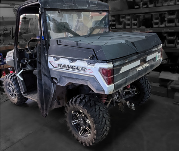 A black Polaris Ranger with a Highlands Ranger 1000/XP1000 (2013-Current) & XP900 (2013-2018) Cargo Box is parked indoors on concrete, cargo bed closed, rugged tires visible, cab door open, and shelves with plastic containers in the background.