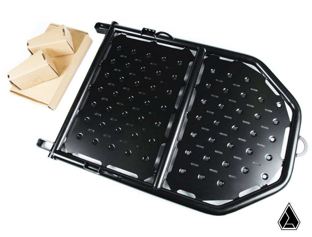 The Assault Industries Adventure Rack (Fits: Polaris RZR XP1K/ Turbo/ Turbo S 14-21) is a black metal folding cargo tray with a perforated surface, shown partially open next to two folded cardboard pieces. A small black geometric logo appears at bottom ri