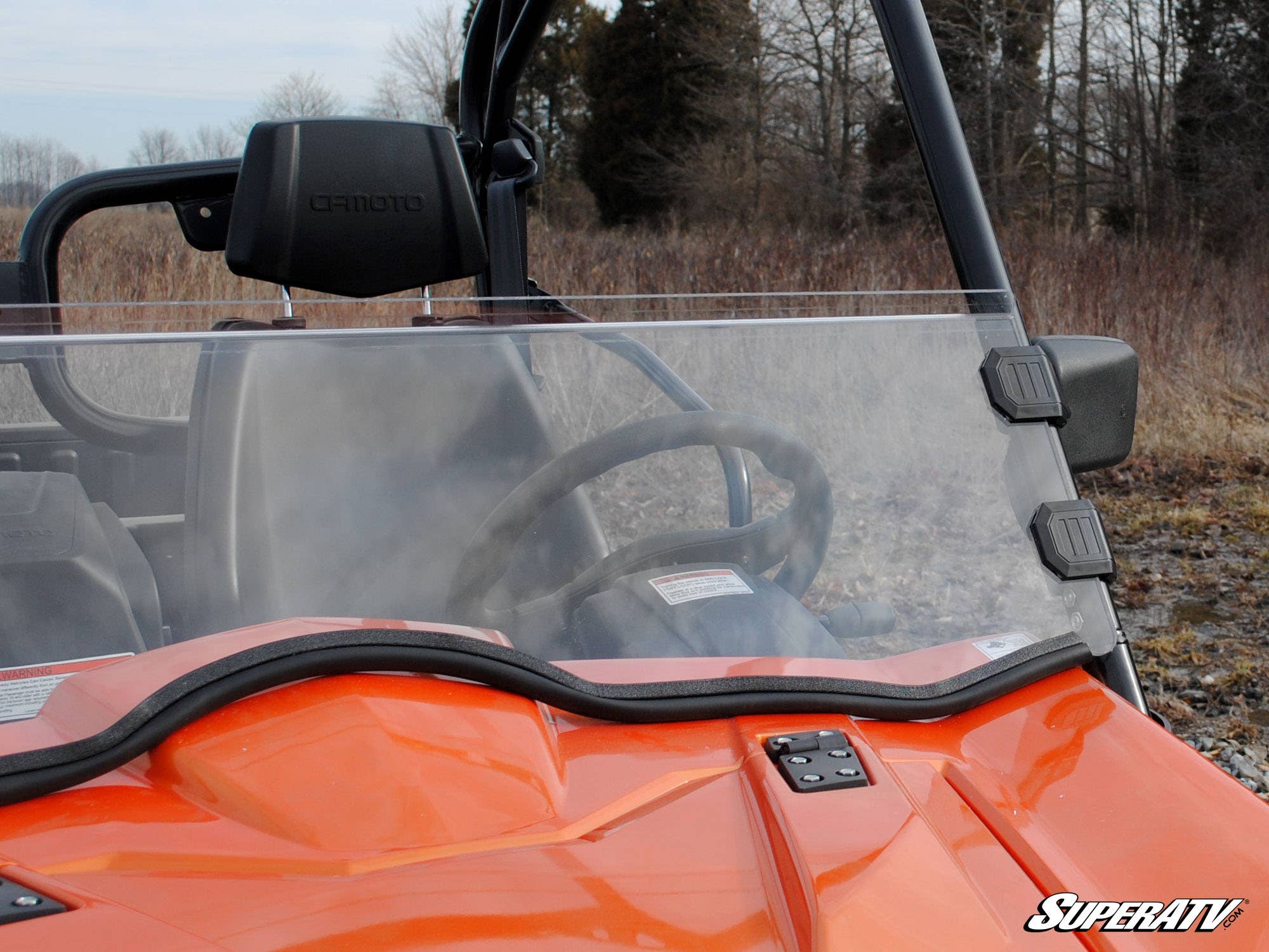 Close-up of a CFMOTO UForce 800 with an orange hood and SuperATVs scratch resistant half windshield attached by black mounts. A clear view of the steering wheel, leafless trees, and dry grass under an overcast sky are visible. SuperATV logo bottom right.