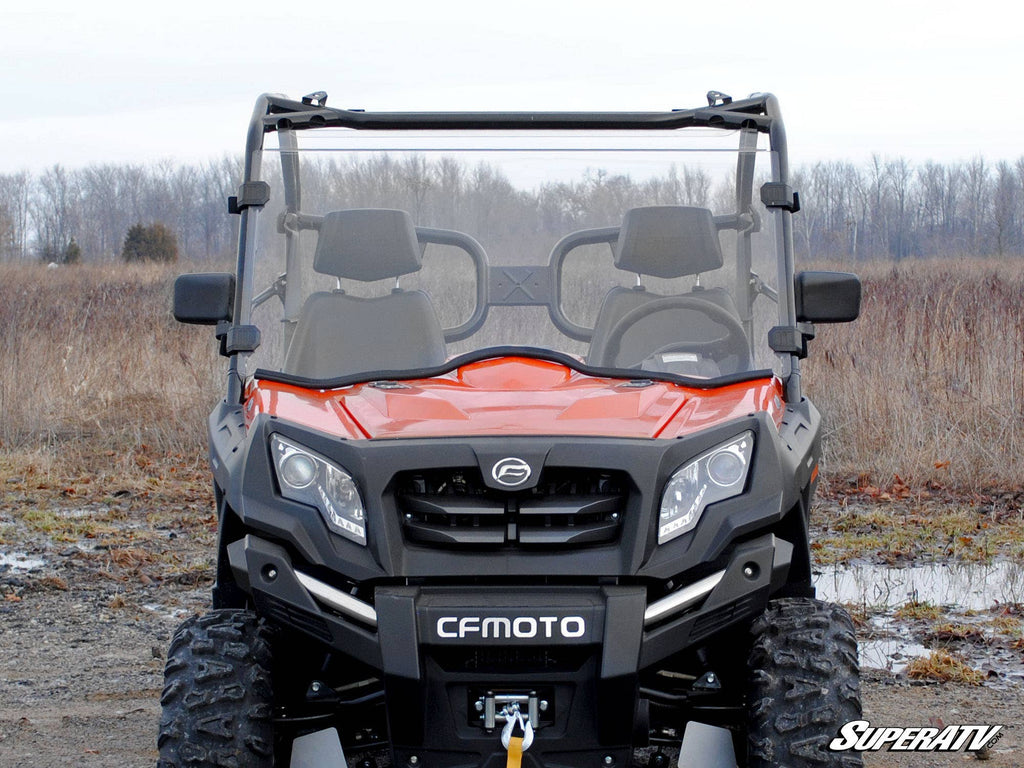 A red CFMOTO UForce 800 with a Scratch-Resistant Full Windshield is parked on a dirt path. Its rugged tires, black grille, and clear windshield stand out. Trees and dry grass appear in the background, with the SuperATV logo at bottom right.