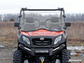 A red CFMOTO UForce 800 with a Scratch-Resistant Full Windshield is parked on a dirt path. Its rugged tires, black grille, and clear windshield stand out. Trees and dry grass appear in the background, with the SuperATV logo at bottom right.
