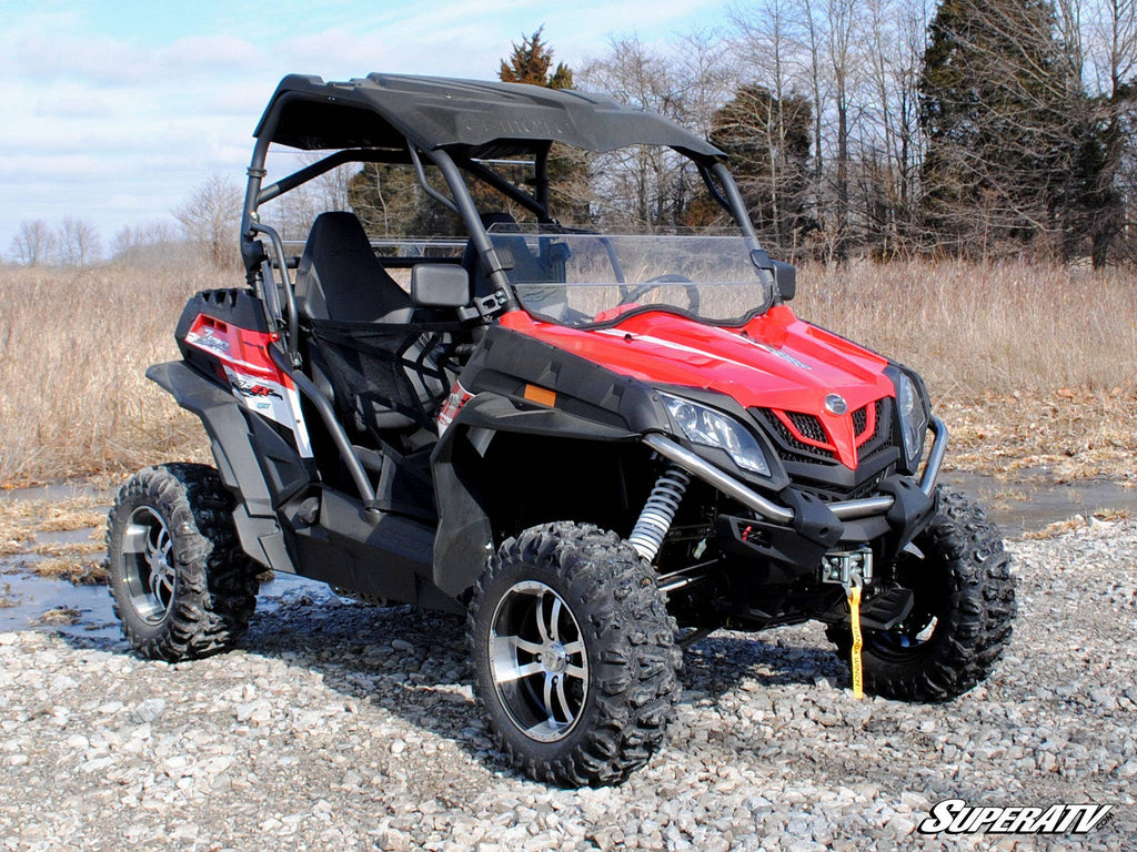 A red and black off-road CFMOTO ZForce 500 with rugged tires, roof, winch, and a Scratch-Resistant Half Windshield sits on rocky ground before dry grass and leafless trees. “SuperATV” is in white text at the lower right of the image.
