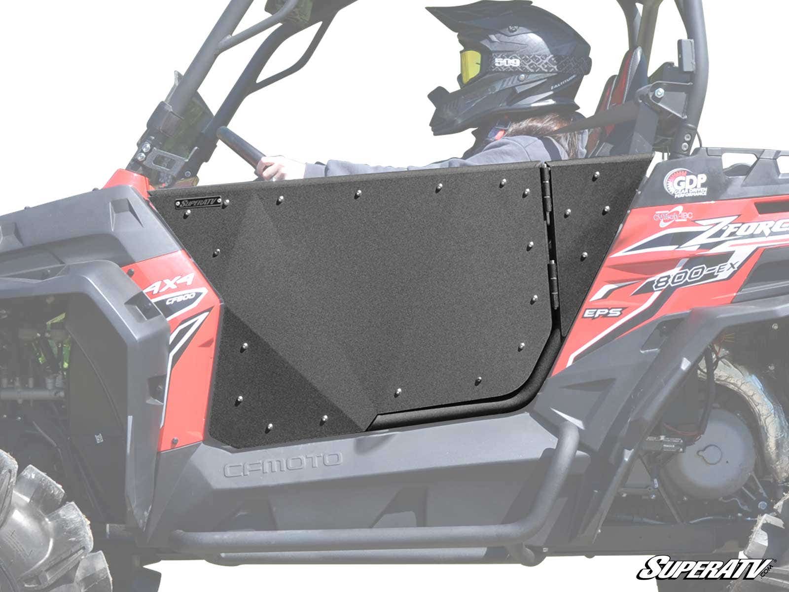 A person wearing a helmet and protective gear sits in a red and black off-road UTV featuring CFMOTO ZForce Aluminum Doors. The SuperATV logo is visible in the bottom right corner.