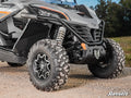 Close-up of the front wheel and suspension system of a rugged vehicle equipped with CFMOTO ZForce 950 Inner Fender Guards. Featuring aggressive tires, visible springs, and a gray body with black details, its parked on a gravel path near grass and trees.