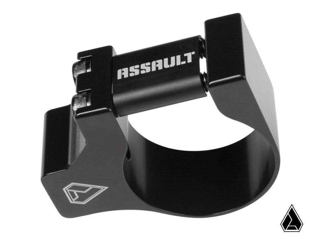 The Assault Industries 1/4-20 Accessory Clamp is a black metal clamp with “ASSAULT” in white on its cylindrical bar, two silver hex bolts, and a white geometric logo on the side. The background is white with a matching logo at the bottom right.