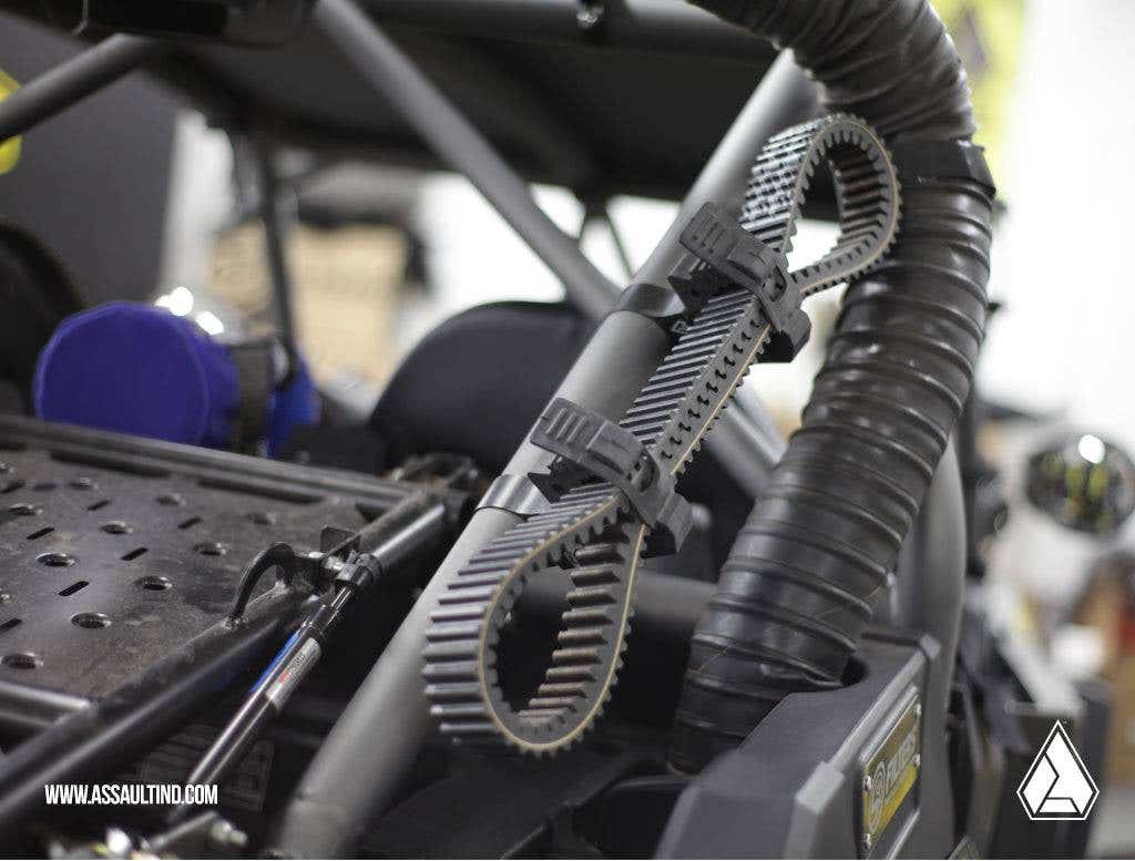 Close-up of a rugged vehicle interior featuring Assault Industries Quickfist Multipurpose Clamps securing a black ribbed belt to the roll cage near a flexible corrugated air duct. ASSAULTIND.COM logo appears in the bottom left corner.