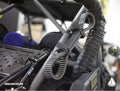 Close-up of a rugged vehicle interior featuring Assault Industries Quickfist Multipurpose Clamps securing a black ribbed belt to the roll cage near a flexible corrugated air duct. ASSAULTIND.COM logo appears in the bottom left corner.