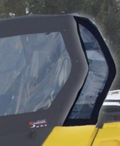 A close-up of Can Am Commander Rear Side Quarter Windows (Set Of 2), black-framed and curved, installed on a yellow vehicle. The transparent windows reveal snow-covered ground and blurred trees in the background.