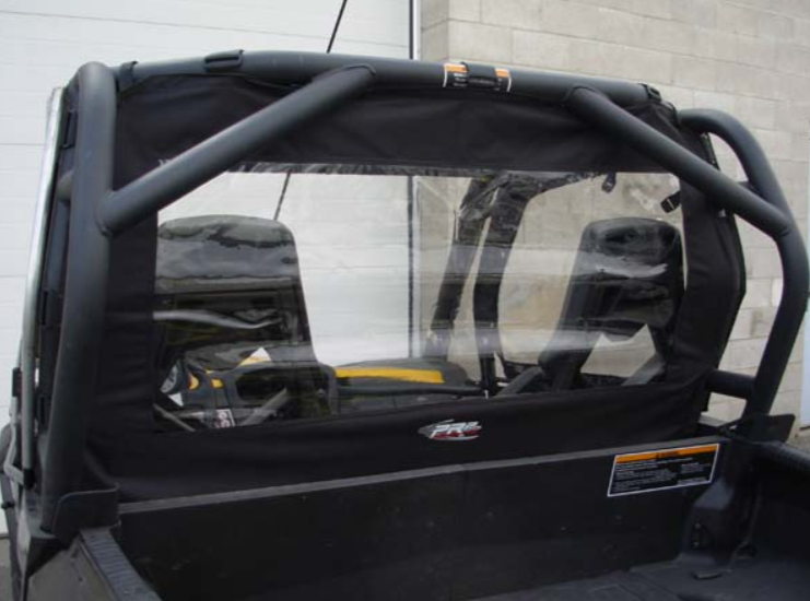 Close-up of a CanAm Commander with Convertible Doors shows a roll cage, two seats, and a clear rear window panel with black border behind the seats, parked near a white overhead door and pale brick wall.