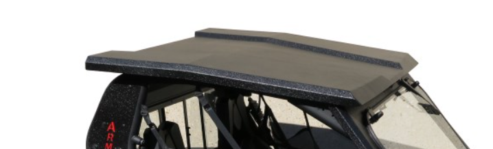 A close-up of the Can-Am Commander Armor Tech Roof, shown in black with a textured finish, angular lines, and a slightly raised center. The roof covers the side-by-side’s passenger area against a white background with some sand visible.