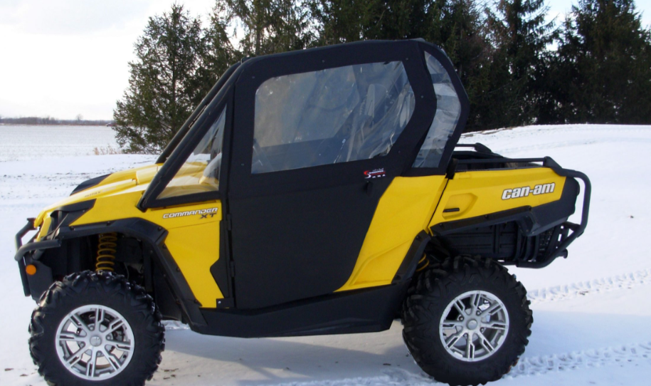 A Can-Am Commander with Convertible Doors, yellow body, black doors and windows, large off-road tires with silver rims, and a roof is parked on snow with evergreen trees and a snow-covered field in the background.