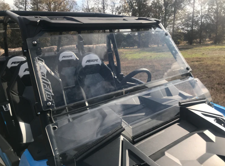 A close-up of a blue side-by-side featuring the RZR XP 1000 Ultimate CoolFlo Windshield with a clear split design and black frame. The interior has two black bucket seats with RZR logos, set against a background of trees and grass in daylight.