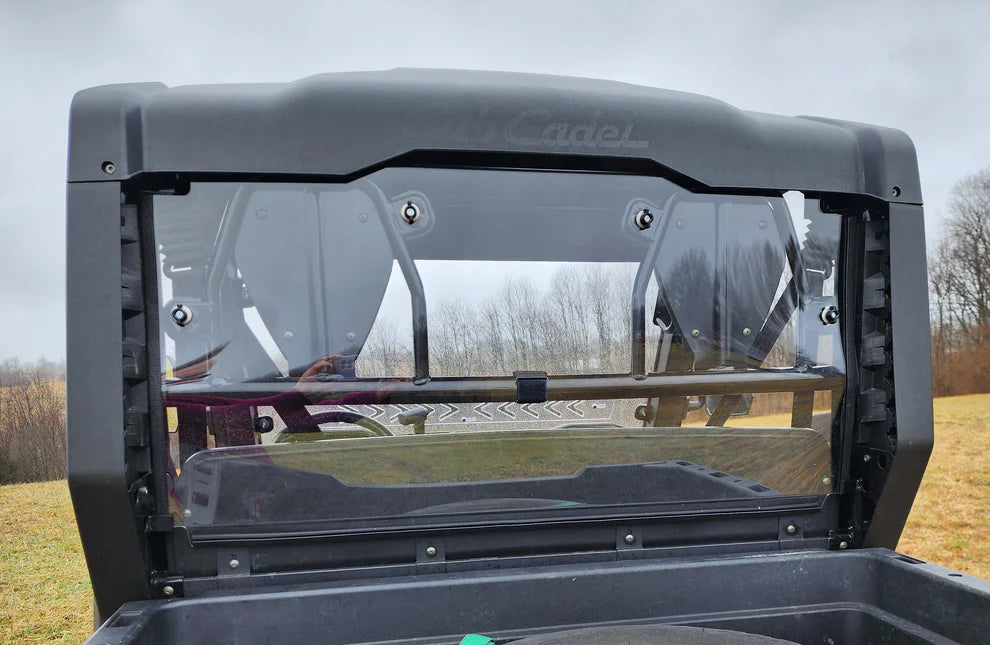 A close-up of the Cub Cadet Challenger MX550/750 Rear Windshield shows the raised black frame and clear plastic window reflecting grass, bare trees, and an overcast sky, with part of the vehicle’s bed and latches visible.