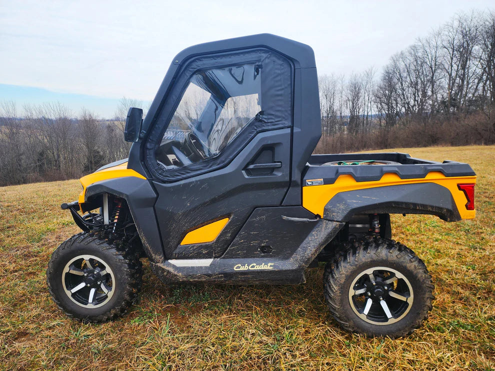 A Cub Cadet Challenger MX550/750 UTV with Upper Doors featuring a steel frame is parked on grass. The yellow and black vehicle has a closed cabin with clear windows, set against leafless trees and a cloudy sky in the background.