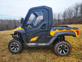 A Cub Cadet Challenger MX550/750 UTV with Upper Doors featuring a steel frame is parked on grass. The yellow and black vehicle has a closed cabin with clear windows, set against leafless trees and a cloudy sky in the background.