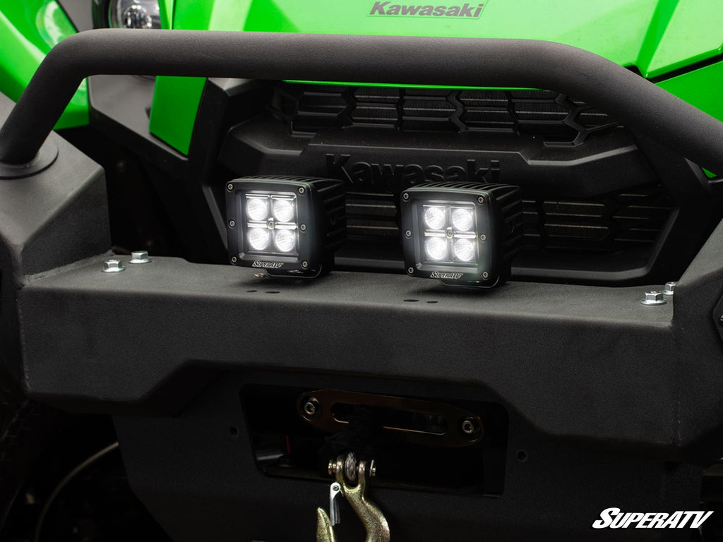 Close-up of a green Kawasaki off-road vehicle with two 3 LED Cube Lights mounted on the black front bumper, a winch and hook below the lights, and the SuperATV logo in the bottom right corner.