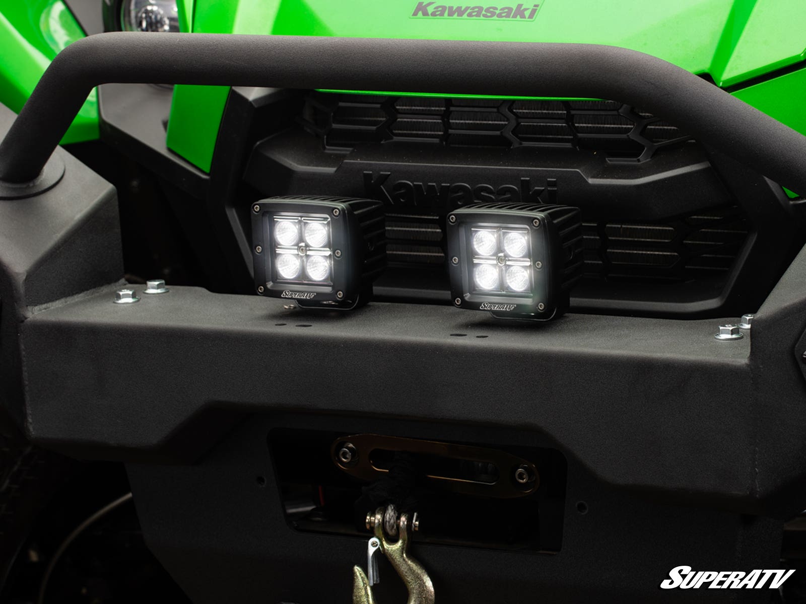 Close-up of a green Kawasaki off-road vehicle with two 3 LED Cube Lights mounted on the black front bumper, a winch and hook below the lights, and the SuperATV logo in the bottom right corner.