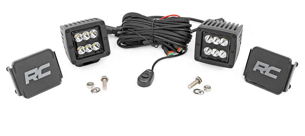 The Defender Game Loader includes two square LED pods (six bulbs each), an RC-labeled wiring harness with switch, mounting covers, and installation hardware—bolts and nuts—all arranged on a white background.