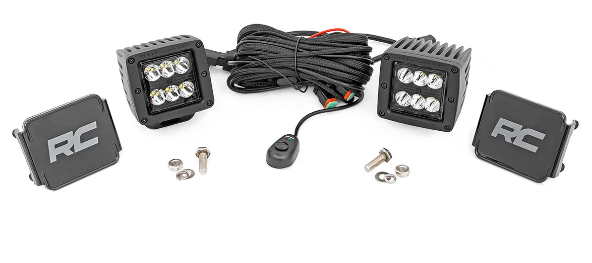 The Defender Game Loader includes two square LED pods (six bulbs each), an RC-labeled wiring harness with switch, mounting covers, and installation hardware—bolts and nuts—all arranged on a white background.